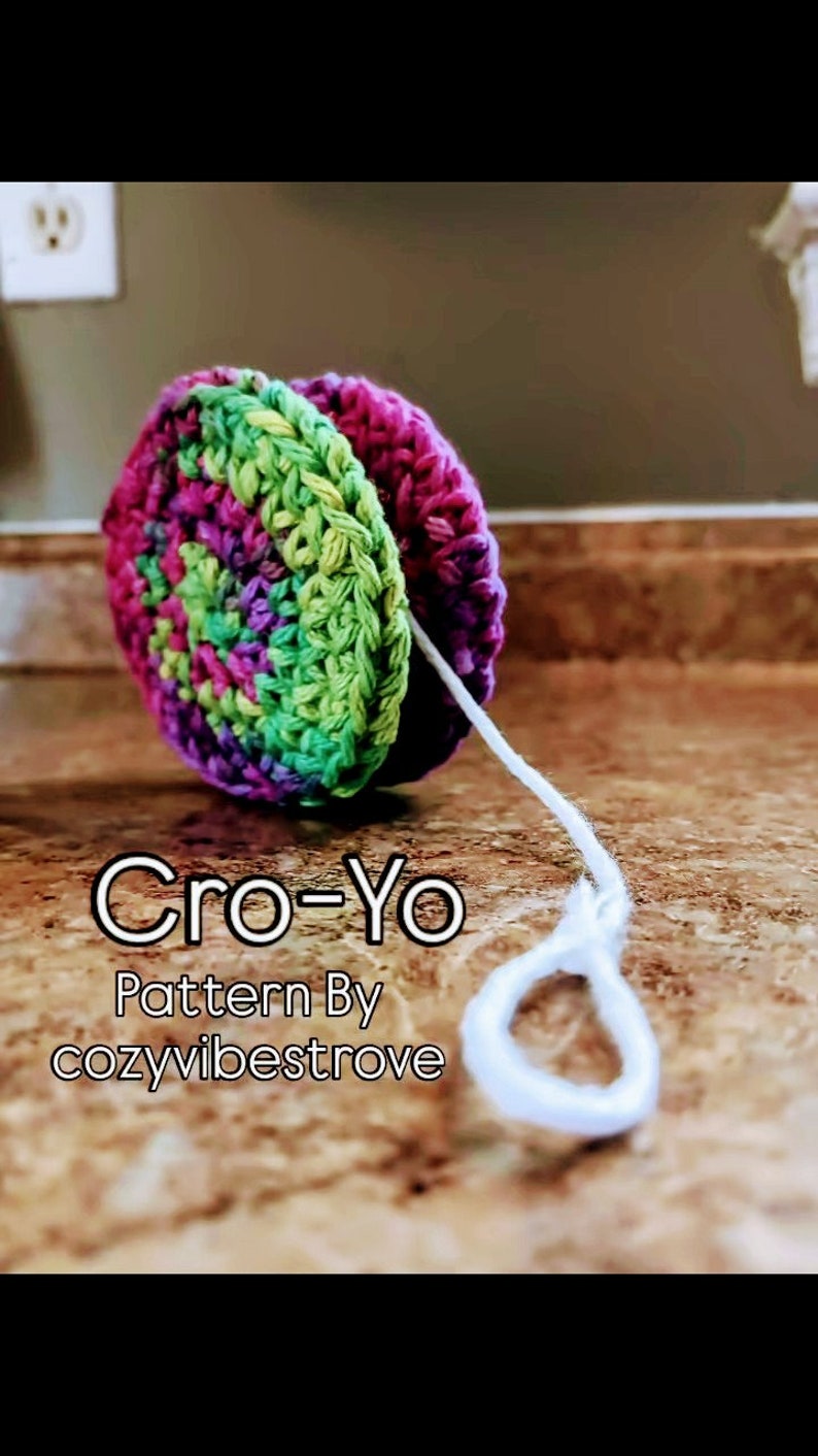 Puede incluir: Un juguete de yoyo de crochet con un esquema de colores verde, morado y rosa. El yoyo est&aacute; hecho con una cuerda blanca. El texto "Cro-Yo Pattern By cozyvibestrove" es visible debajo del yoyo.