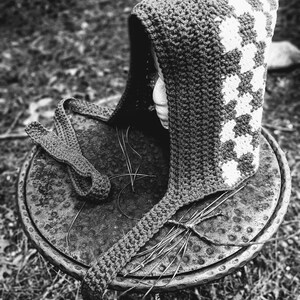 May include: A black and white crocheted hooded hat with a geometric pattern. The hat is lying on a metal table with a textured surface.