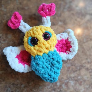 Puede incluir: Un juguete de mariposa de crochet hecho con hilo amarillo, azul, blanco y rosa. La mariposa tiene grandes ojos saltones y un diseño caprichoso.