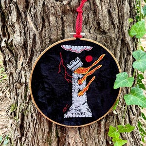 May include: Embroidered hoop art depicting The Tower tarot card. The tower is depicted as a gray structure with flames coming out of the top. The text "The Tower" is embroidered below the tower. The background is black.