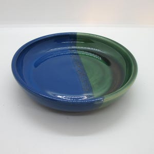 May include: A handmade ceramic bowl with a glossy finish. The bowl is divided into sections of blue, green, and a hint of brown. The round bowl is suitable for serving food or as a decorative piece.