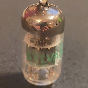 Puede incluir: Un tubo de vacío de vidrio transparente con una parte superior plateada y una etiqueta verde que dice "SYLVANIA" y "MADE IN USA".