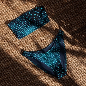 Può includere: Un set bikini a due pezzi composto da un top a fascia senza spalline e uno slip a vita alta. Entrambi i pezzi presentano una base blu navy scuro con un motivo ripetuto di punti e linee ondulate di colore blu ciano brillante.