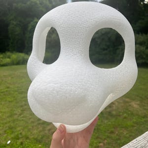 May include: A white, textured, foam-like animal head base for a mask. The structure features a rounded cranium, a prominent snout, and two large, hollowed-out eye sockets. The surface has a porous, granular appearance throughout the entire form.