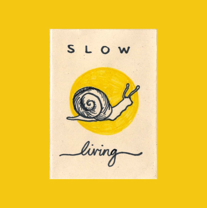 May include: A beige card with black text that reads "SLOW living" and a black and white illustration of a snail crawling on a yellow circle.