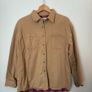 May include: A tan button-down shirt with two chest pockets and a plaid lining. The long-sleeved shirt is hanging on a wooden hanger against a white background. The shirt has a classic, casual style.