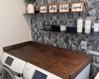 May include: A dark brown wooden countertop sits atop a white washing machine and dryer. The countertop has a lip around the edge. The wall behind the countertop is covered in black and white tropical leaf wallpaper. There are four small wooden signs on the wall that read "wash", "dry", "fold", and "repeat".