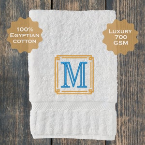 May include: White bath towel with a blue embroidered letter M in a gold frame. The towel is labelled "100% Egyptian Cotton" and "Luxury 700 GSM".