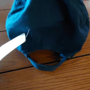 May include: A solid dark teal baseball cap featuring a fabric strap closure at the back with a metal buckle. A white rectangular paper tag is attached to the side of the cap with a plastic fastener. The cap has a button on the top center.