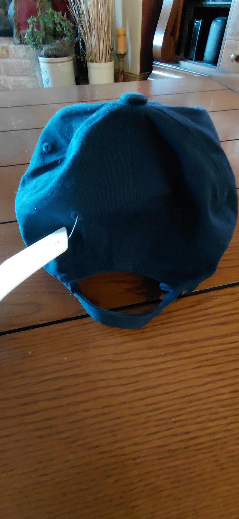 May include: A solid dark teal baseball cap featuring a fabric strap closure at the back with a metal buckle. A white rectangular paper tag is attached to the side of the cap with a plastic fastener. The cap has a button on the top center.