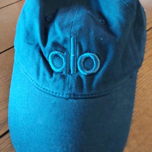 May include: A solid black baseball cap featuring a structured six-panel design with a curved brim and a fabric-covered button at the top. The word alo is embroidered in a matching black thread on the front center of the cap crown.