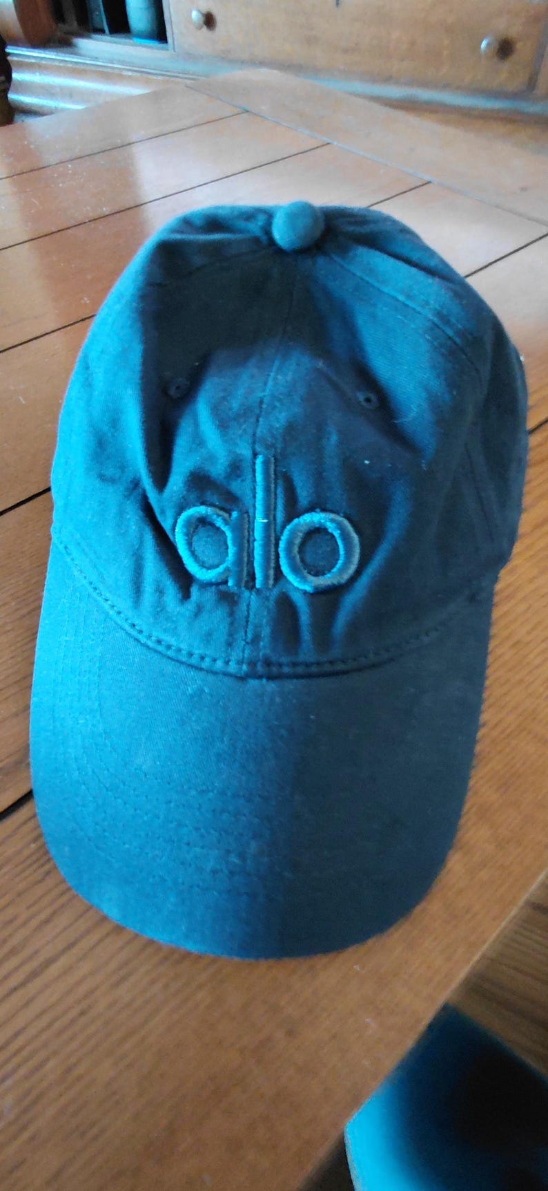 May include: A solid black baseball cap featuring a structured six-panel design with a curved brim and a fabric-covered button at the top. The word alo is embroidered in a matching black thread on the front center of the cap crown.
