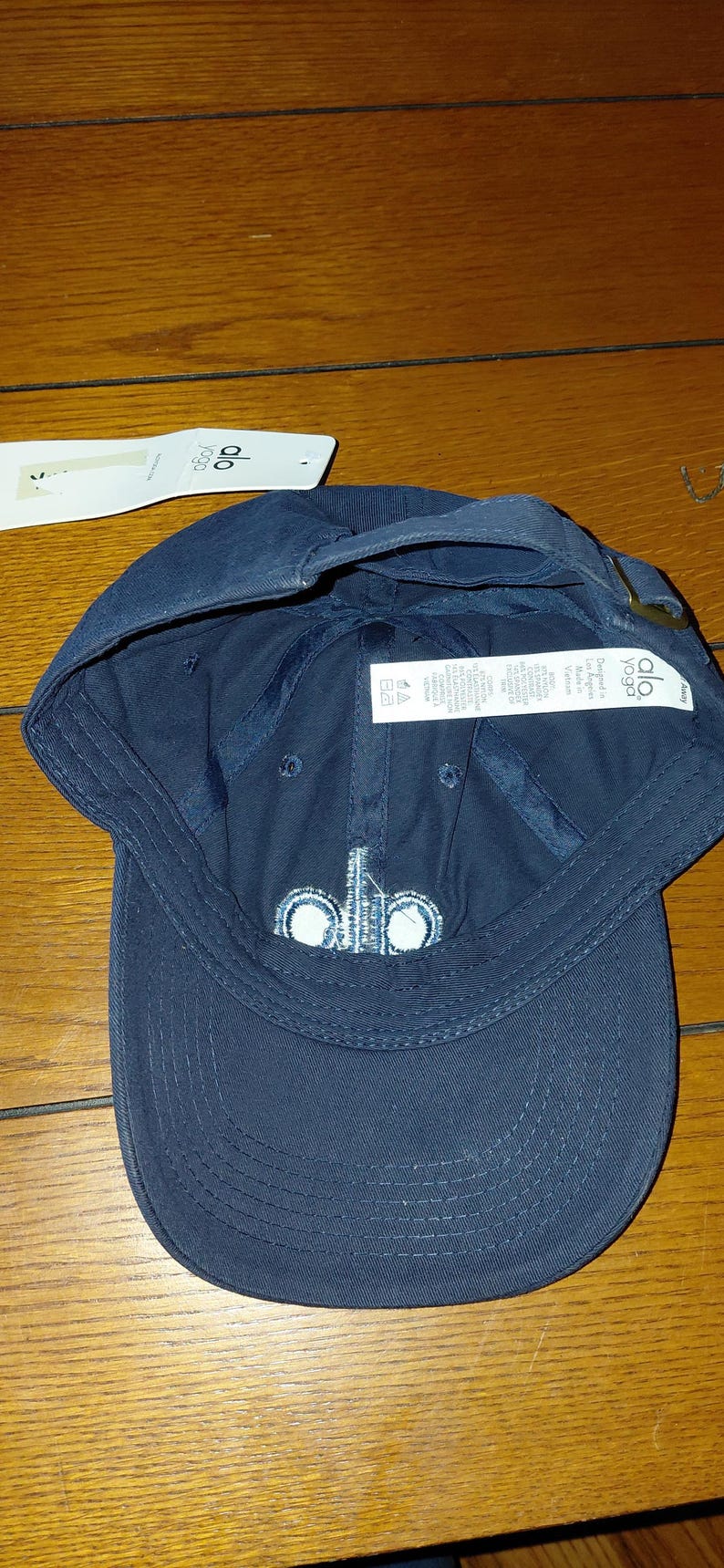 May include: A navy blue baseball cap featuring the white embroidered text alo on the front panel. The cap includes a white paper hang tag with the brand logo and an interior white fabric care label with text detailing material composition.