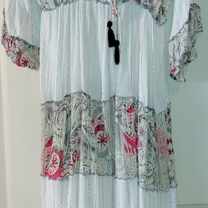May include: White dress with a V-neck and long sleeves. The dress features a tiered design with patterned fabric in shades of pink, gray, and white. Silver metallic threads run vertically throughout the dress, adding a subtle shimmer. A black tassel hangs from the neckline.