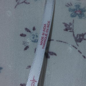 Op de afbeelding: Close-up van een witte stof met een bloemenpatroon in blauw en paars. Een label leest "MADE IN INDIA / FABRIQUE EN INDE" en toont maat "L".