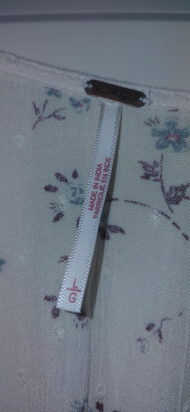 Op de afbeelding: Close-up van een witte stof met een bloemenpatroon in blauw en paars. Een label leest "MADE IN INDIA / FABRIQUE EN INDE" en toont maat "L".
