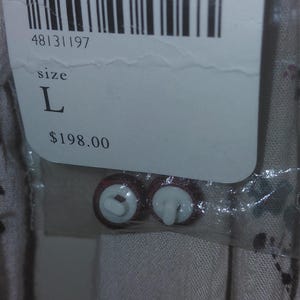 Op de afbeelding: Een productlabel met informatie, waaronder maat "L" en de kleur "IVRY/IVOIR". Twee bruine en witte knopen zijn zichtbaar. Het label bevat een barcode en de tekst "FL_5".