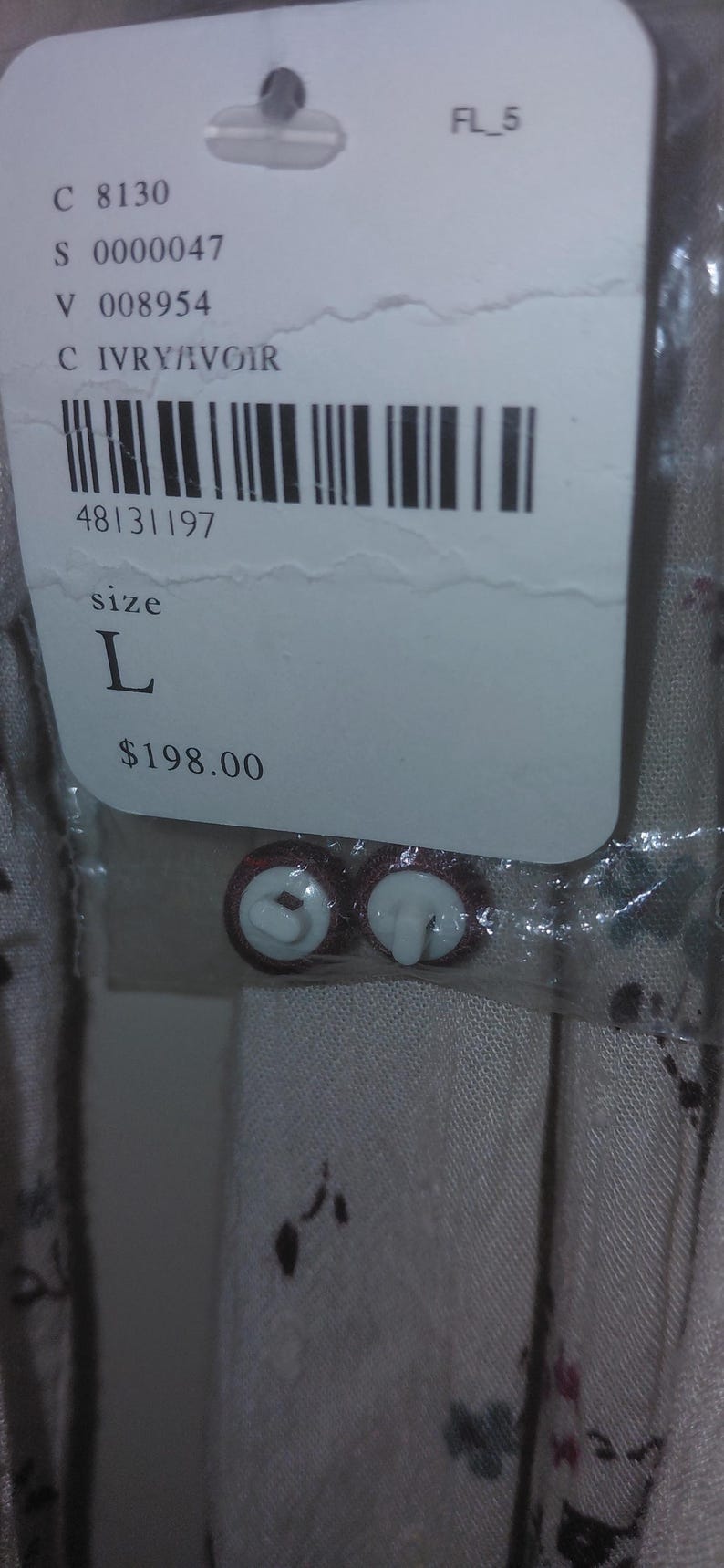 Op de afbeelding: Een productlabel met informatie, waaronder maat "L" en de kleur "IVRY/IVOIR". Twee bruine en witte knopen zijn zichtbaar. Het label bevat een barcode en de tekst "FL_5".