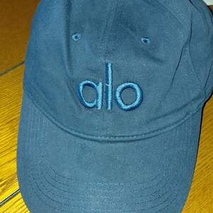 May include: A dark blue baseball cap featuring the word alo embroidered in a matching thread color on the front panel. The cap has a curved brim with visible stitching and a fabric-covered button at the top center of the crown.