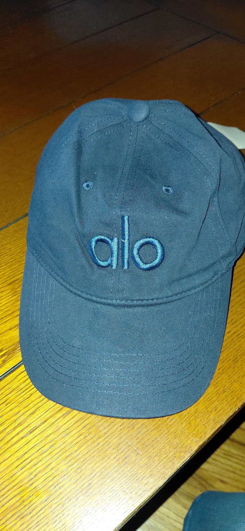 May include: A dark blue baseball cap featuring the word alo embroidered in a matching thread color on the front panel. The cap has a curved brim with visible stitching and a fabric-covered button at the top center of the crown.