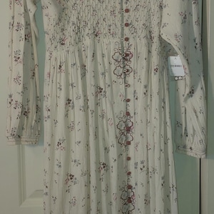 Op de afbeelding: Cr&egrave;mekleurige lange jurk met een bloemenpatroon in roze en paars. Het heeft lange mouwen, een V-hals en een knoopsluiting aan de voorkant. De bodice heeft een gerimpeld detail. De jurk hangt aan een zwarte hanger.