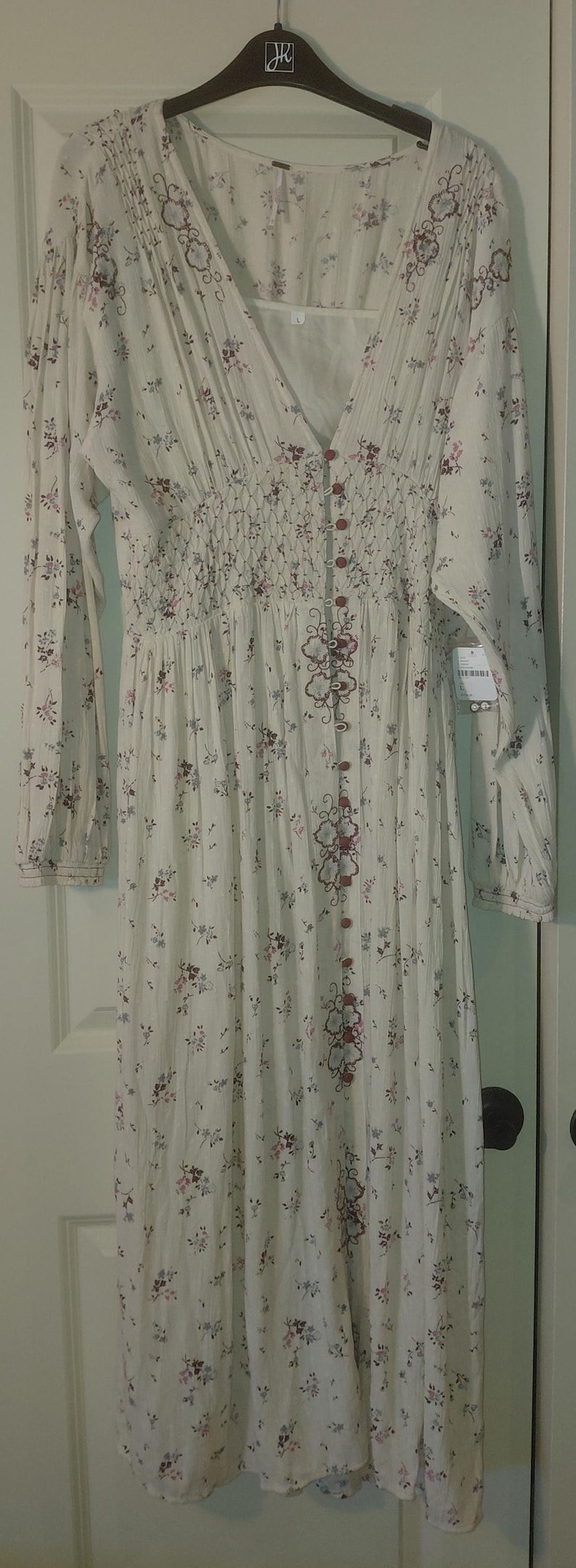 Op de afbeelding: Cr&egrave;mekleurige lange jurk met een bloemenpatroon in roze en paars. Het heeft lange mouwen, een V-hals en een knoopsluiting aan de voorkant. De bodice heeft een gerimpeld detail. De jurk hangt aan een zwarte hanger.