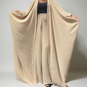 May include: A beige, flowing outfit consisting of a long, open-front robe and matching wide-leg pants. The robe has long sleeves and drapes elegantly. The pants have a high waist with a knot detail. The outfit is made of a textured fabric.