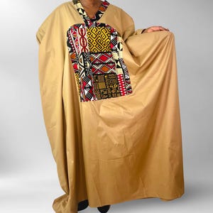 Puede incluir: Un caftán beige con cuello en V y un bolsillo grande y colorido. El bolsillo presenta un patrón geométrico en rojo, negro, amarillo y blanco. El escote tiene un patrón contrastante en blanco y negro.