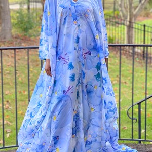 Könnte beinhalten: Ein langes, fließendes Kleid mit V-Ausschnitt und langen Ärmeln. Das Kleid ist hellblau mit einem Blumenmuster in Blau- und Weißtönen. Es hat eine Knopfleiste vorne und einen bodenlangen Rock.