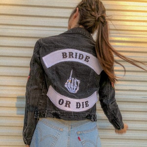 Puede incluir: Una chaqueta vaquera negra con un parche blanco que dice "Bride or Die" y una mano de esqueleto con un anillo en el dedo. La chaqueta tiene un aspecto desgastado y la lleva puesta una persona con el pelo largo y castaño.