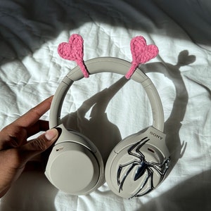 May include: Beige wireless headphones with a silver spider design on one earcup. The headphones have pink crocheted hearts attached to the headband.