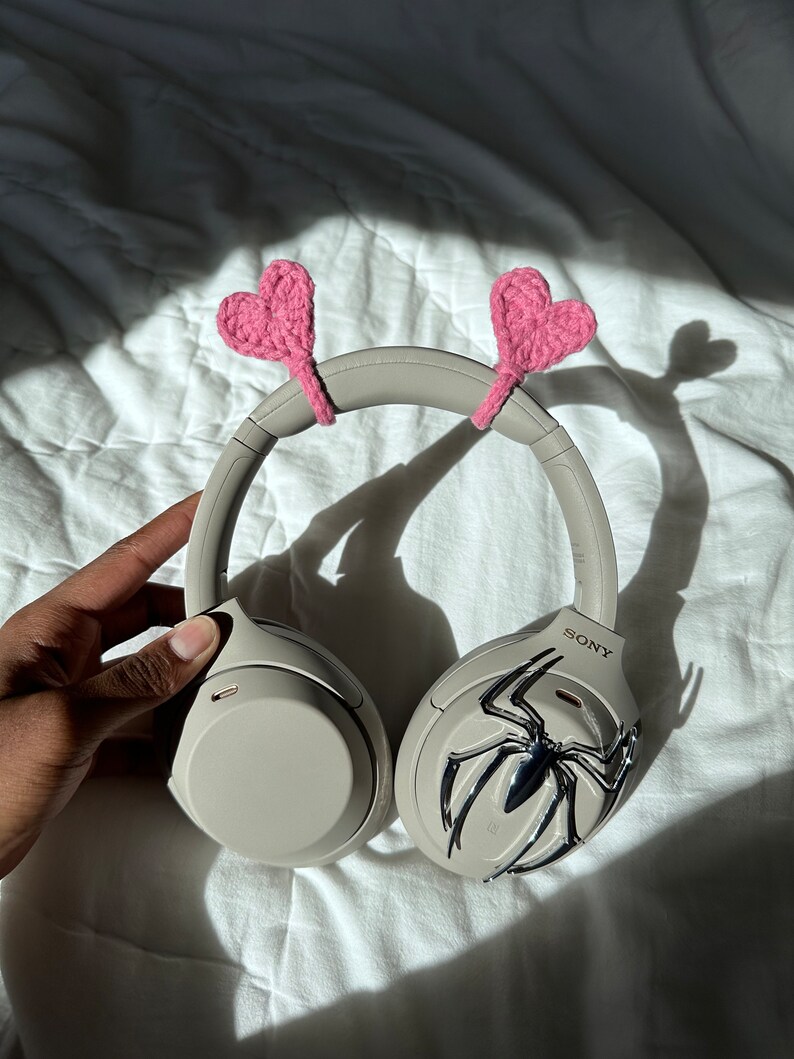 May include: Beige wireless headphones with a silver spider design on one earcup. The headphones have pink crocheted hearts attached to the headband.