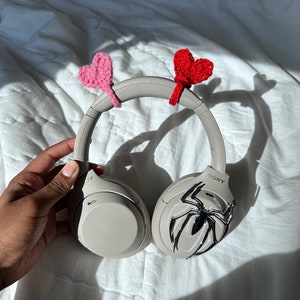 May include: Gray over-ear headphones with a spider-man design on one earcup. The headphones have pink and red crocheted hearts attached to the headband.
