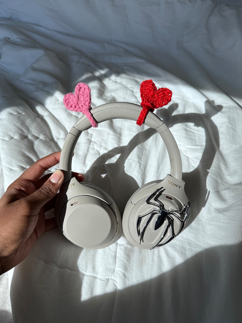 May include: Gray over-ear headphones with a spider-man design on one earcup. The headphones have pink and red crocheted hearts attached to the headband.