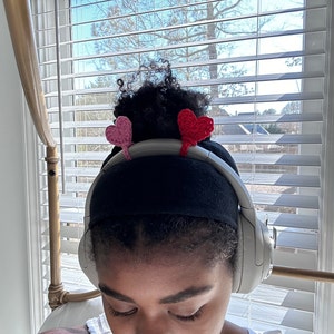 May include: A person wearing black headphones with pink and red heart-shaped crocheted embellishments on the headband.