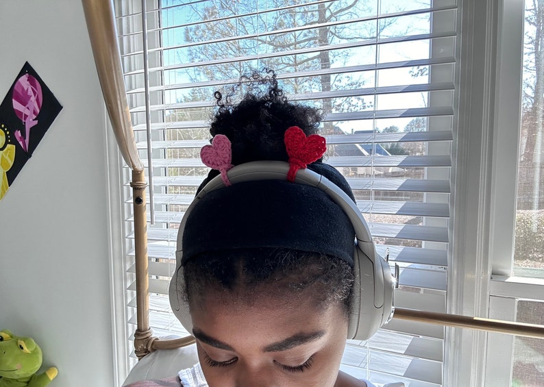 May include: A person wearing black headphones with pink and red heart-shaped crocheted embellishments on the headband.