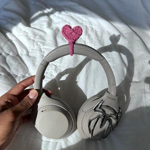 May include: A pair of gray noise-canceling headphones with a pink crocheted heart attached to the headband. The headphones have a silver spider design on one earcup and the word "SONY" on the other.