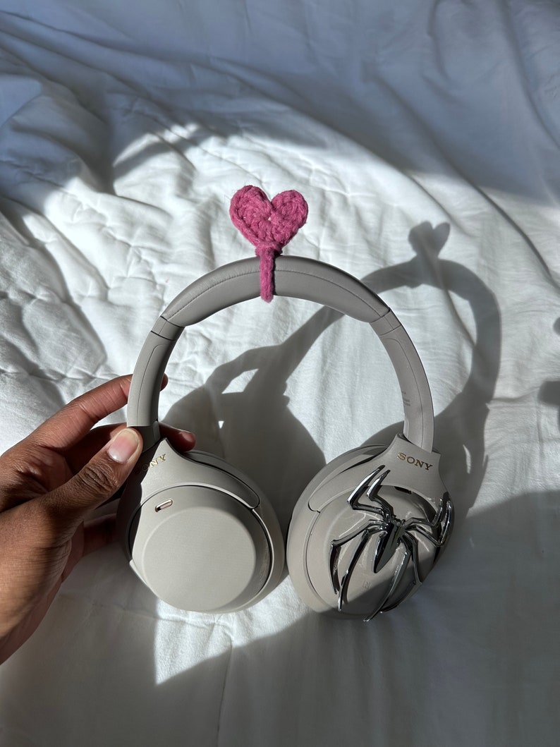 May include: A pair of gray noise-canceling headphones with a pink crocheted heart attached to the headband. The headphones have a silver spider design on one earcup and the word "SONY" on the other.