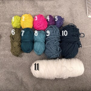 May include: Eleven balls of yarn in various colors, including green, yellow, pink, purple, blue, and white. The yarn is arranged in two rows, with the larger white ball of yarn in the bottom row.