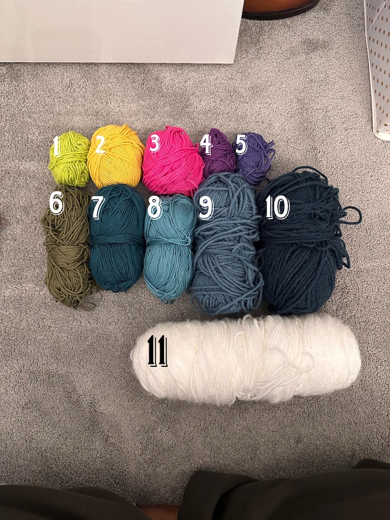 May include: Eleven balls of yarn in various colors, including green, yellow, pink, purple, blue, and white. The yarn is arranged in two rows, with the larger white ball of yarn in the bottom row.