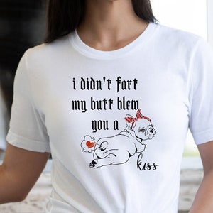 Op de afbeelding: Wit T-shirt met de tekst "i didn't fart my butt blew you a kiss" en een afbeelding van een hond met een rode strik en hart. Het shirt wordt gecombineerd met een spijkerbroek short.