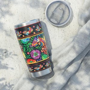 May include: A stainless steel travel tumbler with a black background and a vibrant floral and bird design. The colorful pattern includes flowers, birds, and foliage in shades of red, yellow, blue, and green. The lid is off to the side.