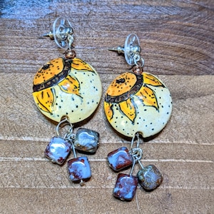 May include: A pair of dangle earrings with a white ceramic disc painted with a yellow sunflower design. The earrings have a silver hook and dangle with three blue and brown glass beads.