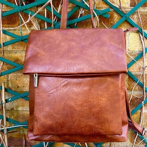 May include: A brown faux leather backpack with a flap closure and a silver zip. The bag has a textured finish and adjustable straps. The background features a green trellis and a brick wall.