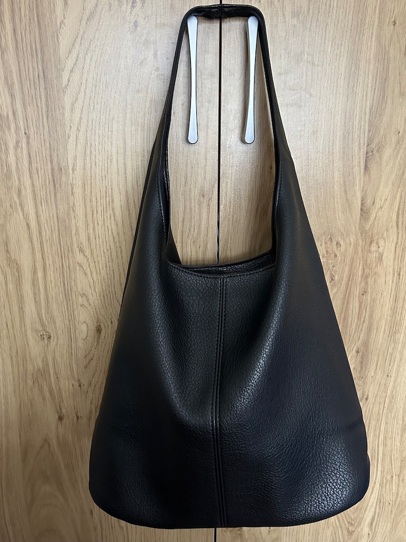 May include: A black leather shoulder bag with a textured surface hangs from a silver handle. The bag has a simple, minimalist design and a large capacity. The bag is made of a durable material and is suitable for everyday use.