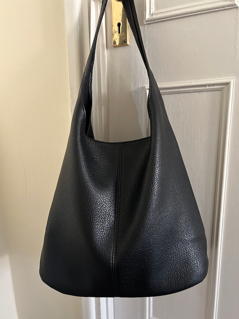 May include: A black leather hobo bag with a textured surface. The bag has a single shoulder strap and a spacious interior. The bag is hanging on a door.