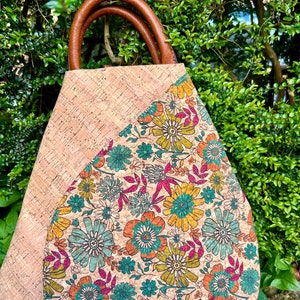 Può includere: Una borsa tote in sughero con manici in pelle marrone. La borsa presenta un motivo floreale nei toni del giallo, verde acqua e rosa su sfondo beige. La borsa è ambientata su uno sfondo di fogliame verde.