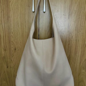 May include: A light beige shoulder bag with a rounded bottom and long straps. The bag is hanging on a wooden door with silver handles. The bag has a simple, minimalist design.