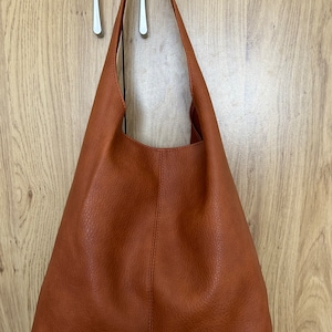 May include: A brown leather shoulder bag with a single strap. The bag has a simple, minimalist design with a textured surface and a stitched seam down the center. The bag is hanging against a light wood-grain background.
