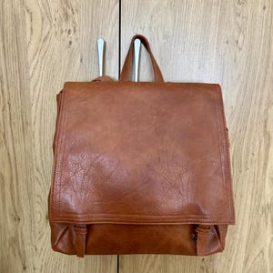 May include: A medium-sized, cognac-coloured backpack with a large, rectangular fold-over flap. The bag features a textured, leather-like finish, a top handle, and two vertical straps with buckle-style closures at the bottom of the front panel.